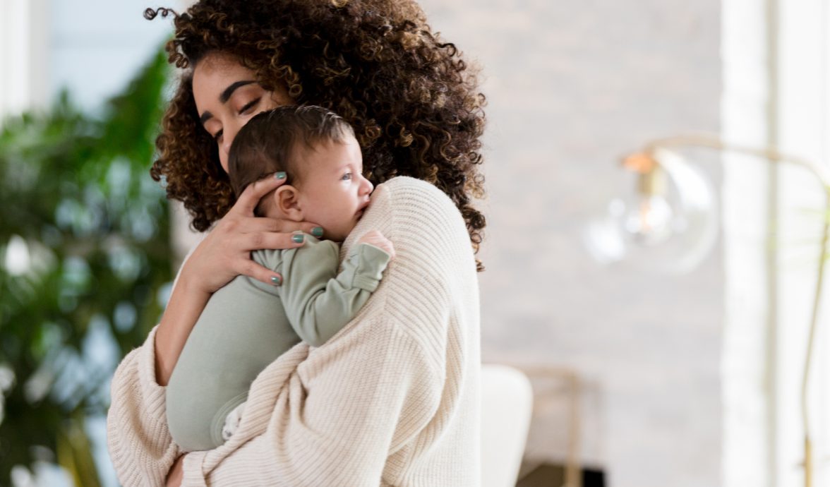 Imagem de mãe em pé com bebê no colo, uma das formas de acalmar a cólica em recém-nascido.