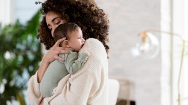 Imagem de mãe em pé com bebê no colo, uma das formas de acalmar a cólica em recém-nascido.