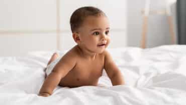 Imagem de um bebê de bruços em cima de um lençol branco, em cima da cama. Ele está apenas de fralda, tem a pele morena, olhos e cabelos escuros e está sorrindo.