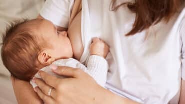 Imagem focada em um bebê no seio da mãe, alimentado-se com leite materno. Ele tem a pele clara e o cabelo castanho claro. Está com um body branco e o olhar em direção a mãe, que veste uma camiseta branca.