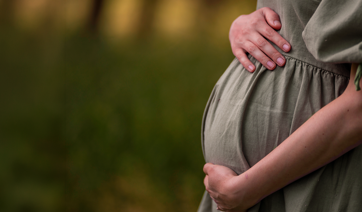 Imagem da barriga de uma gestante. Ela está com as mãos sobre a barriga e usa um vestido verde oliva.