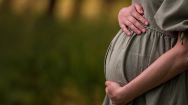 Imagem da barriga de uma gestante. Ela está com as mãos sobre a barriga e usa um vestido verde oliva.