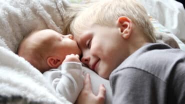 Imagem de um bebê e uma criança. Ambos tem a pele e o cabelo claros e estão deitados. O irmão mais velho está abraçando o bebê.