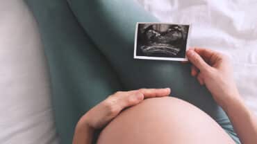 Imagem fechada da barriga de uma gestante. Ela está sentada, com uma das mãos na barriga e a outra segurando uma imagem de ultrassom, exame realizado para identificar o percentil fetal do bebê.