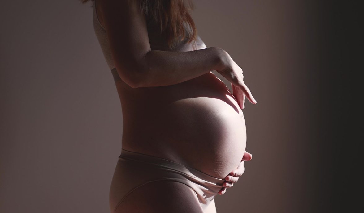 Imagem do corpo de uma gestante. Ela está de perfil e as suas mãos envolvem a barriga. A imagem está escura com um feixe de luz incidindo sobre a barriga.
