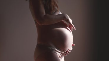 Imagem do corpo de uma gestante. Ela está de perfil e as suas mãos envolvem a barriga. A imagem está escura com um feixe de luz incidindo sobre a barriga.