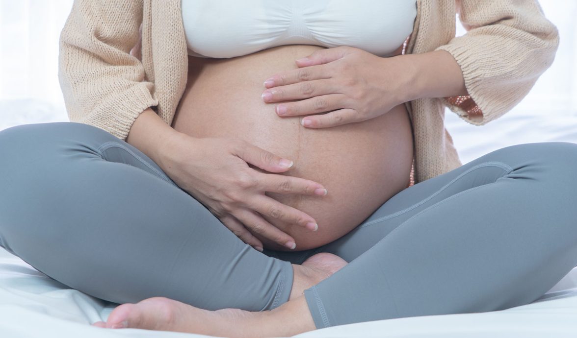 Imagem do corpo de uma gestante. Ela tem a pele clara, veste um top branco, uma caça de fginástica cinza e um cardigan creme. Está com as pernas em posição de borboleta e acariciando a barriga com as mãos.