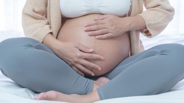 Imagem do corpo de uma gestante. Ela tem a pele clara, veste um top branco, uma caça de fginástica cinza e um cardigan creme. Está com as pernas em posição de borboleta e acariciando a barriga com as mãos.