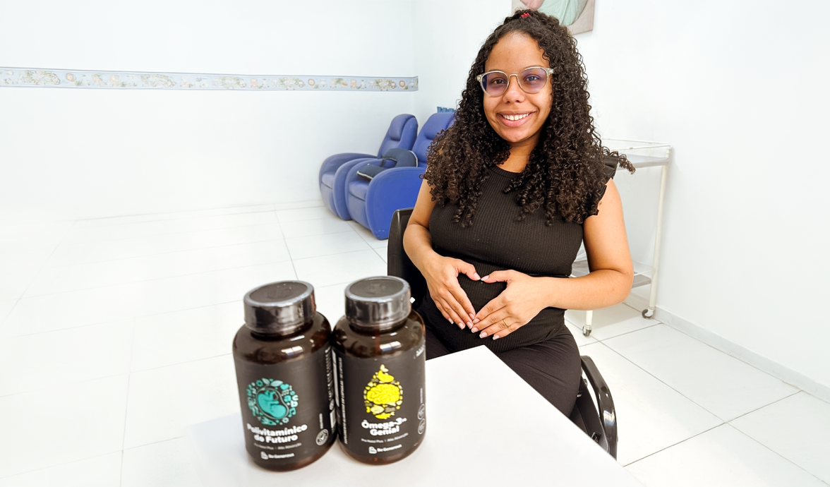 Imagem de uma gestante sentada, fazendo um coração com as mãos sobre a barriga. Na mesa a sua frente estão os suplementos doados pelo projeto Be Generous em São Gonçalo, RJ.