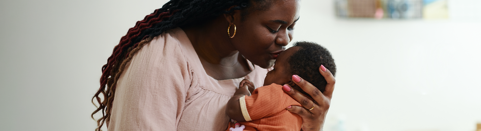 Imagem de uma mulher dando colo para o bebê. Ela tem a pele negra, os cabelos escuros, encaracolados e longos. Está de olhos fechados e dando um beijo na testa do bebê, que também tem a pele negra e veste um body laranja.
