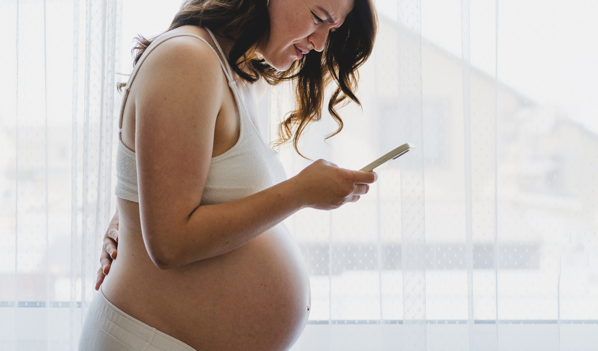 Imagem de uma mulher grávida. Ela está com um celular em uma das mãos e a outra nas costas, com expressão de dor, um indicativo de que a bolsa rompeu.