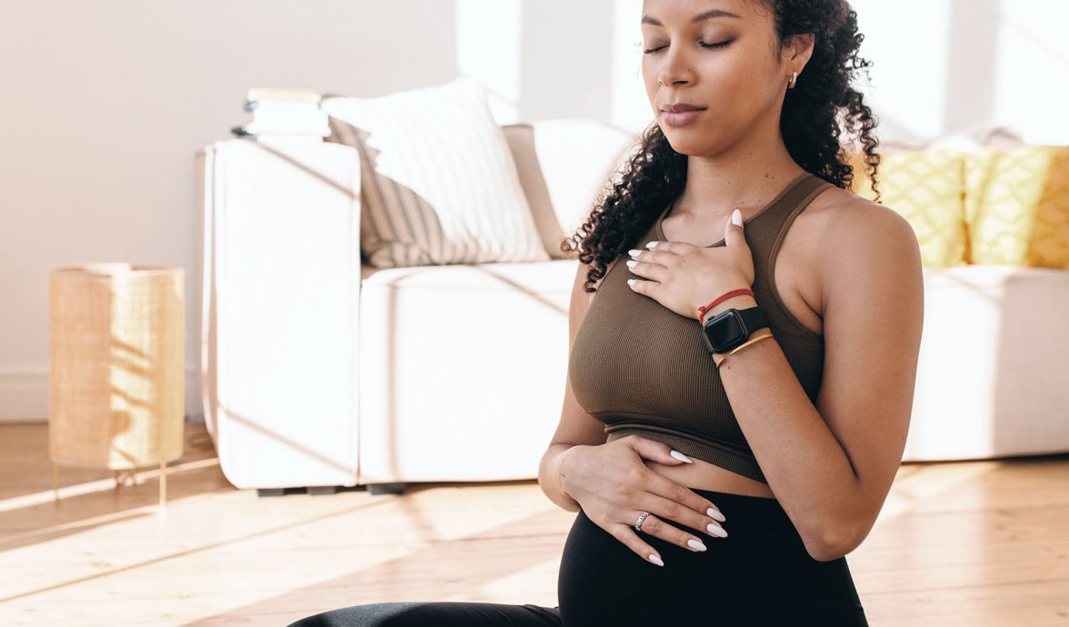 Imagem de uma gestante sentada no chão, com os olhos fechados, uma mão sobre a barriga e outra no peito. Ela está com as pernas cruzadas e controlando a respiração, uma das técnicas utilizadas para aliviar a dor do parto.