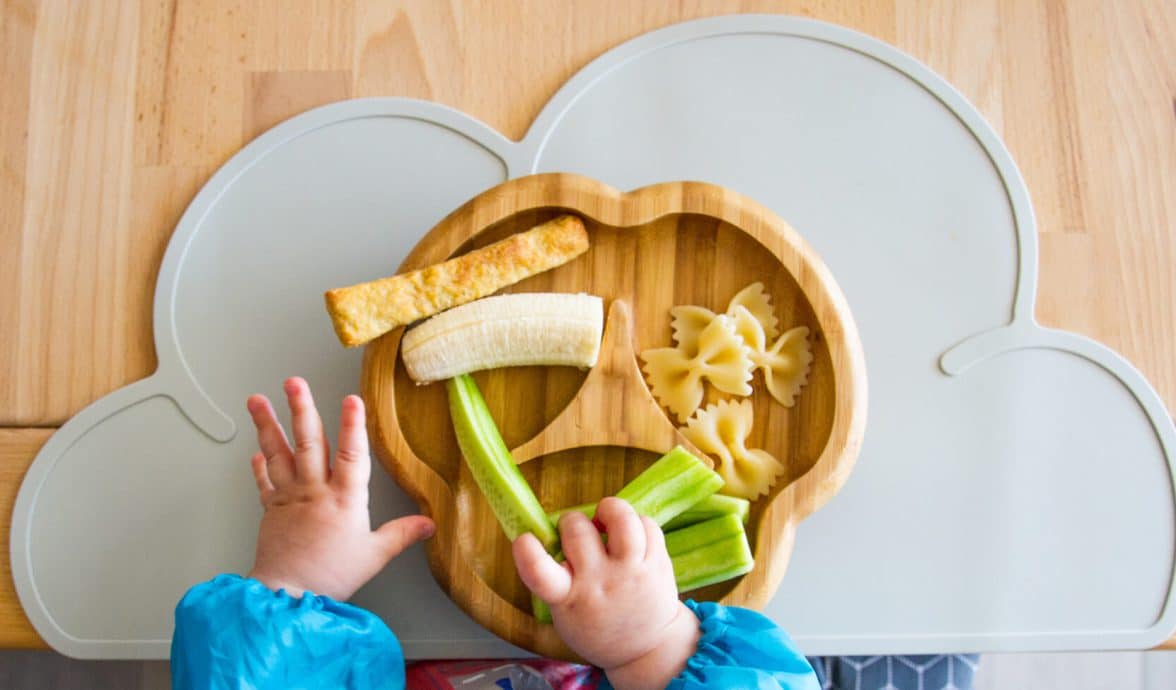 mãos de um bebê adepto ao BLW seguram alimentos em pedaços sob o pratinho de comida.
