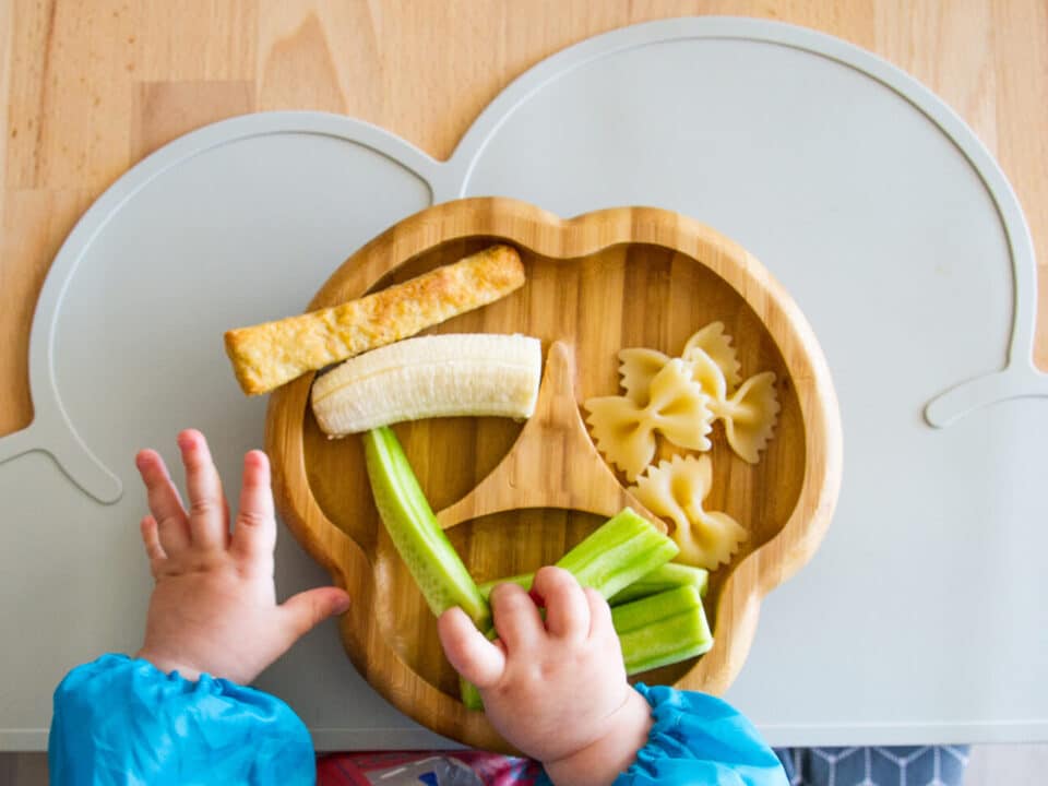 mãos de um bebê adepto ao BLW seguram alimentos em pedaços sob o pratinho de comida.