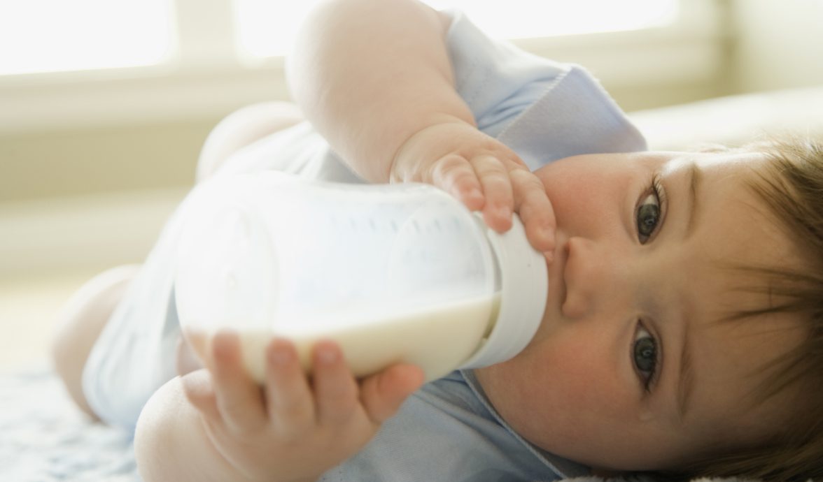 Imagem fechada de bebê mamando. Ele está segurando a mamadeira com as duas mãos e com olhar voltdado para a câmera.