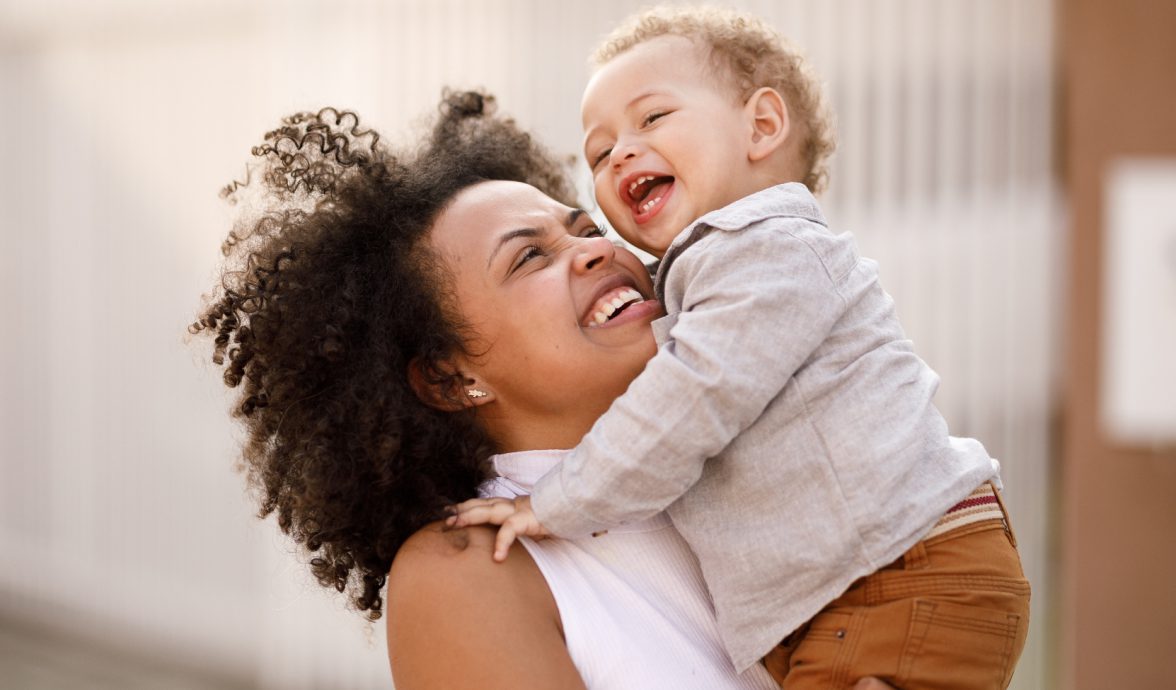 Mãe em pé com criança pequena no colo. A mãe está olhando para o bebê e ambos estão sorrindo, uma relação de maternidade real.