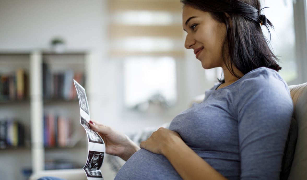 Gestante sentada com a mão sobre a barriga, olhando imagens de ultrassom do bebê, um dos exames obrigatórios no pré-natal.