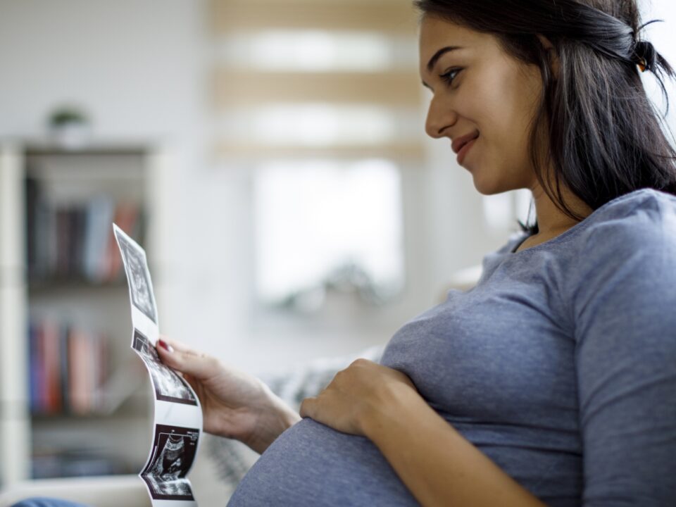 Gestante sentada com a mão sobre a barriga, olhando imagens de ultrassom do bebê, um dos exames obrigatórios no pré-natal.