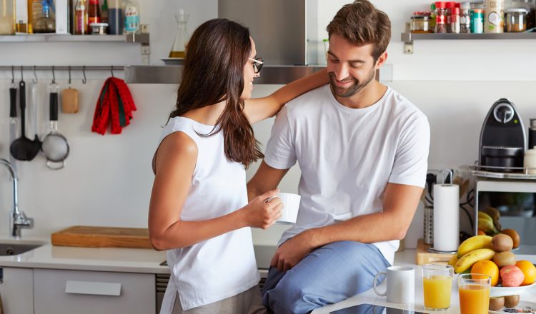 Imagem de um casal na cozinha. A mulher está segurança uma xícara na mão direita, olhando para ele e com a mão esquerda em volta do seu pescoço. Ele está sentado sobre a bancada e olhando em direção a barriga dela. Em cima da bancada está uma bandeja com várias furtas, dois copos de suco e um xícara. Ambos estão sorrindo.