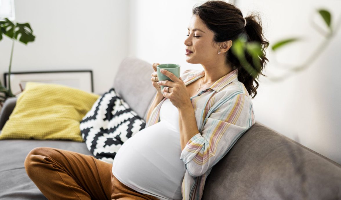 Imagem de uma gestante sentada no sofá, com as pernas entrelaçadas. Ela está segurando uma xícara de chá com as duas mãos e com os olhos fechados, como se esse alimento lhe causasse uma sensação de bem-estar.