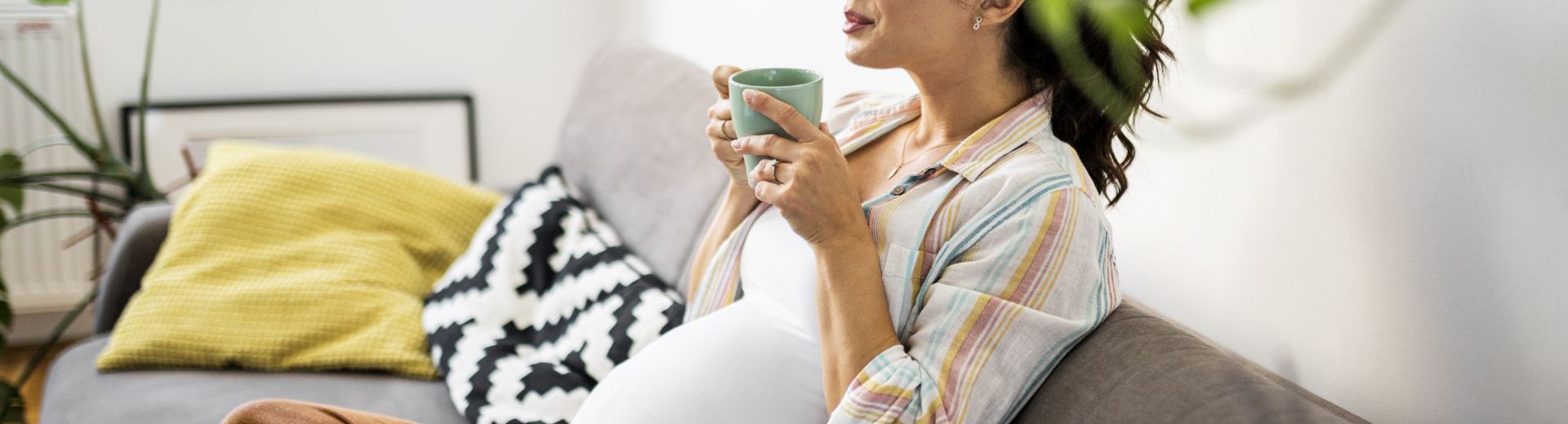 Imagem de uma gestante sentada no sofá, com as pernas entrelaçadas. Ela está segurando uma xícara de chá com as duas mãos e com os olhos fechados, como se esse alimento lhe causasse uma sensação de bem-estar.