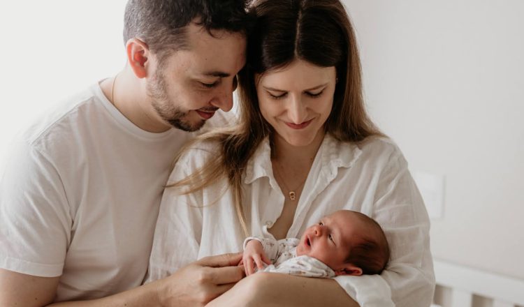 Imagem de um casal com um bebê. A mãe está como bebê nos braços. Ela está olhando em direção ao rostinho dele e sorrindo. O pai está com um dos braços nas costas da mãe e o outro segurando o bebê. Ele também está olhando em direção ao bebê e sorrindo.