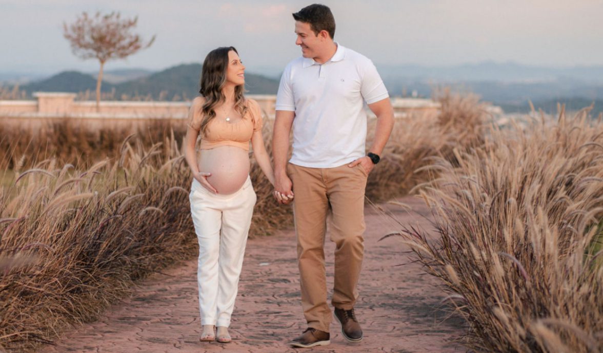 Imagem de uma casal andando de mãos dadas. Eles estão sorrindo e olhando um para o outro. A mulher está grávida e segura a barriga com a mão direita. O homem está com a mão no bolso da calça.