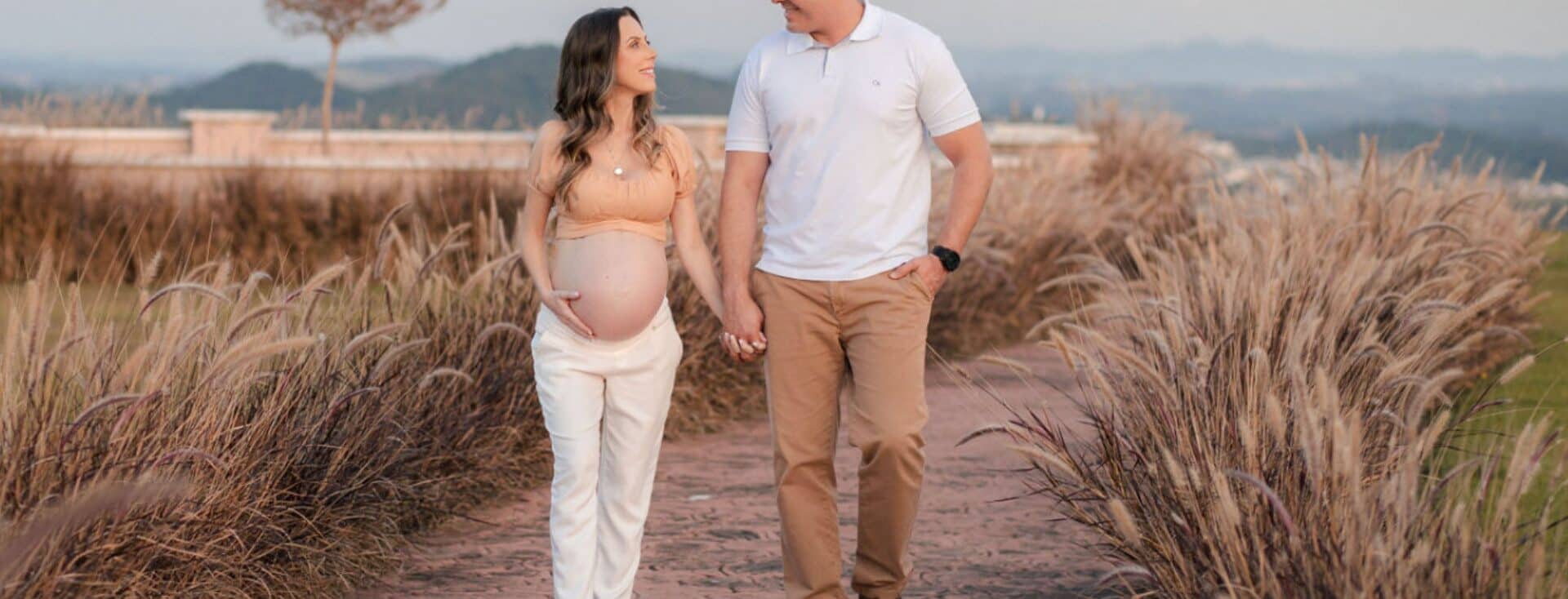Imagem de uma casal andando de mãos dadas. Eles estão sorrindo e olhando um para o outro. A mulher está grávida e segura a barriga com a mão direita. O homem está com a mão no bolso da calça.