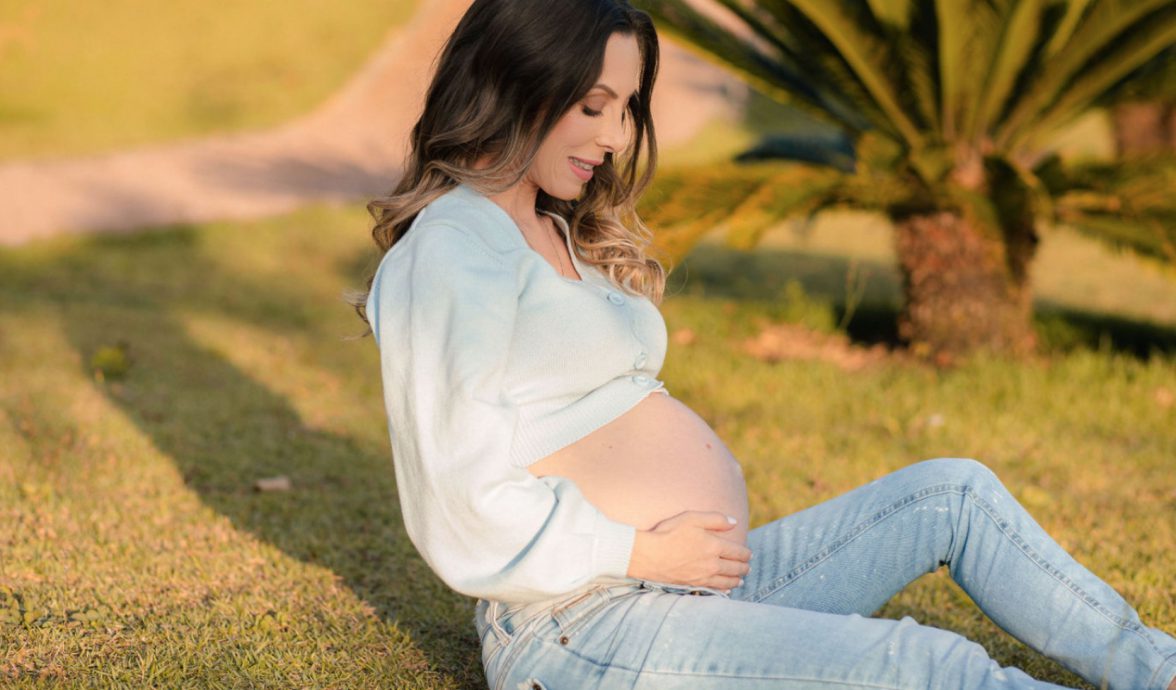 Imagem de uma gestante sentada na grama. Ela veste uma calça jeans e uma blusa de linho azul bebê, mostrando a barriga. Ela está olhando em direção à barriga. A mão esquerda está apoiada no chão e a mão direita segura a barriga.