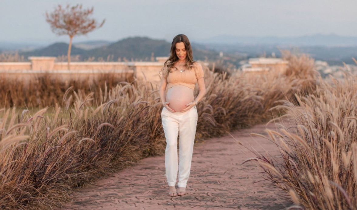 Imagem de uma gestante andando ao ar livre e segurando a barriga com as mãos. Seu olhar está direcionado para a barriga. Ela está vestindo um top caramelo e uma calça de alfaiataria creme.