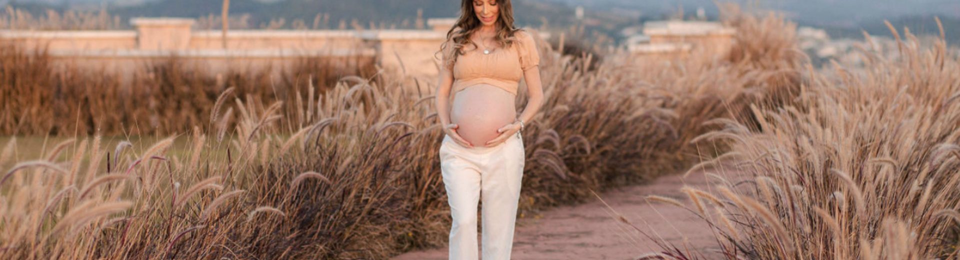 Imagem de uma gestante andando ao ar livre e segurando a barriga com as mãos. Seu olhar está direcionado para a barriga. Ela está vestindo um top caramelo e uma calça de alfaiataria creme.