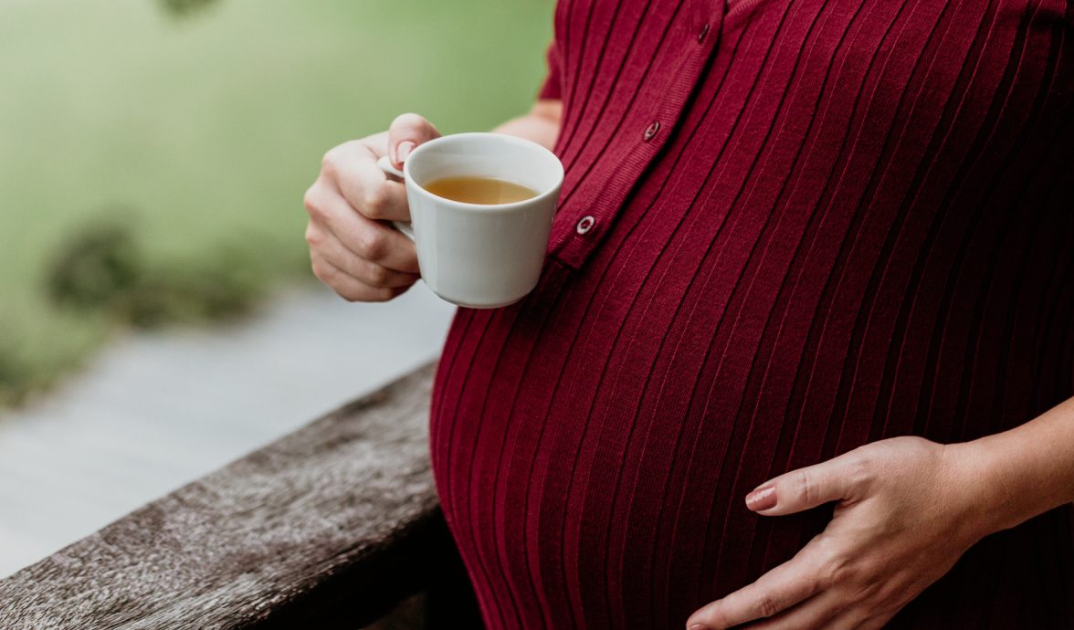 Imagem de uma gestante tomando chá na gravidez. A imagem mostra apenas a barriga e as mãos, sendo que a mão direita está segurando a xícara de chá e a esquerda está na barriga. A gestante usa uma blusa na cor marsala.