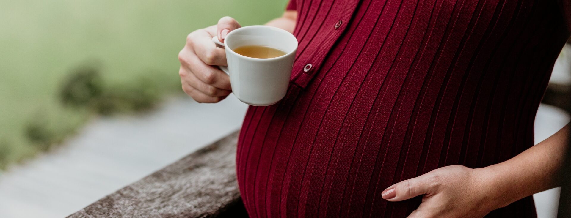 Imagem de uma gestante tomando chá na gravidez. A imagem mostra apenas a barriga e as mãos, sendo que a mão direita está segurando a xícara de chá e a esquerda está na barriga. A gestante usa uma blusa na cor marsala.