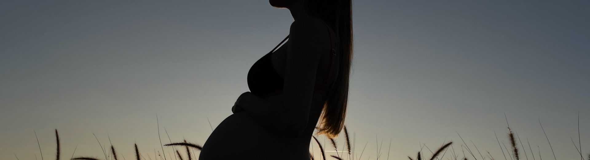 Uma gestante no segundo trimestre de gestação, contra a luz do sol em um campo aberto, com a mão na barriga.