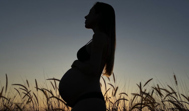 Uma gestante no segundo trimestre de gestação, contra a luz do sol em um campo aberto, com a mão na barriga.