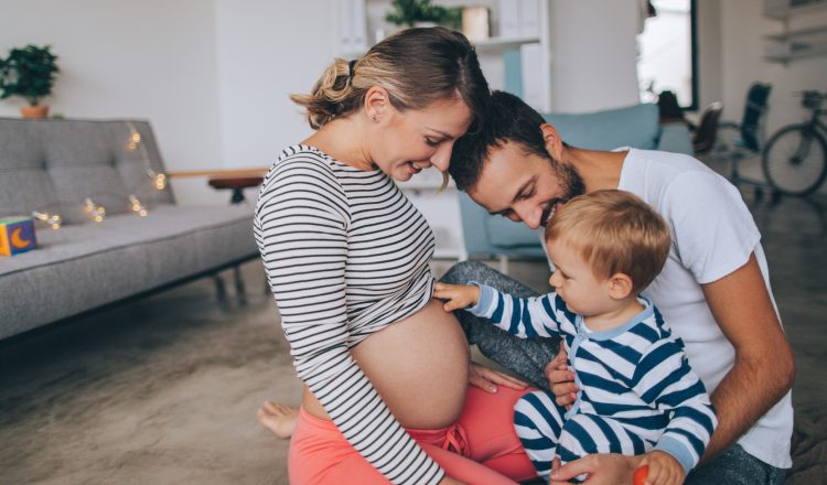 Imagem de uma família. Pai, mãe e um menino de mais ou menos dois anos estão juntos, sorrindo. A mãe está grávida e o menino está com a mão na barriga, estimulando o bebê.
