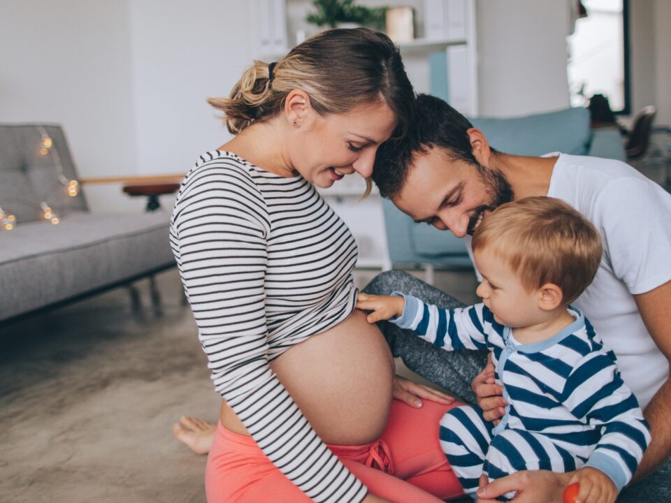 Imagem de uma família. Pai, mãe e um menino de mais ou menos dois anos estão juntos, sorrindo. A mãe está grávida e o menino está com a mão na barriga, estimulando o bebê.