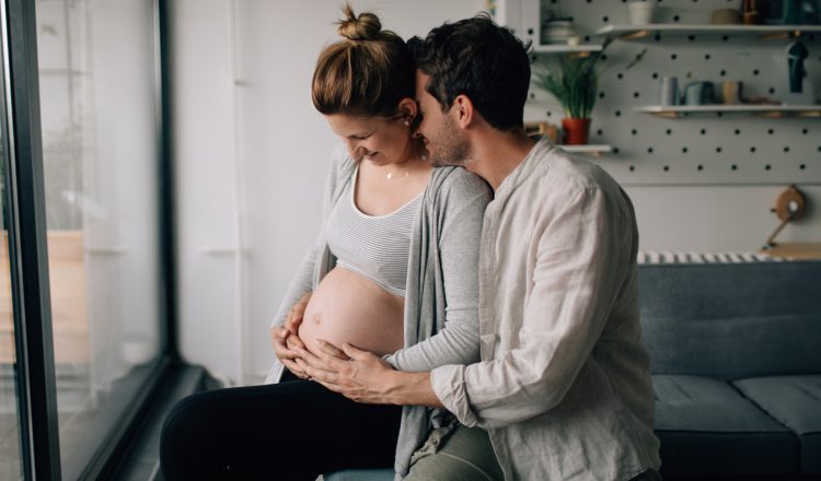 Imagem de um casal. Eles estão na sala de casa. Ela está grávida e parece estar se preparando para o parto. Ele está com os braços no entorno da barriga dela e com o rosto encostado em sua nuca, enquanto ela olha para a barriga e sorri. Ambos estão com as mãos no pé da barriga dela.