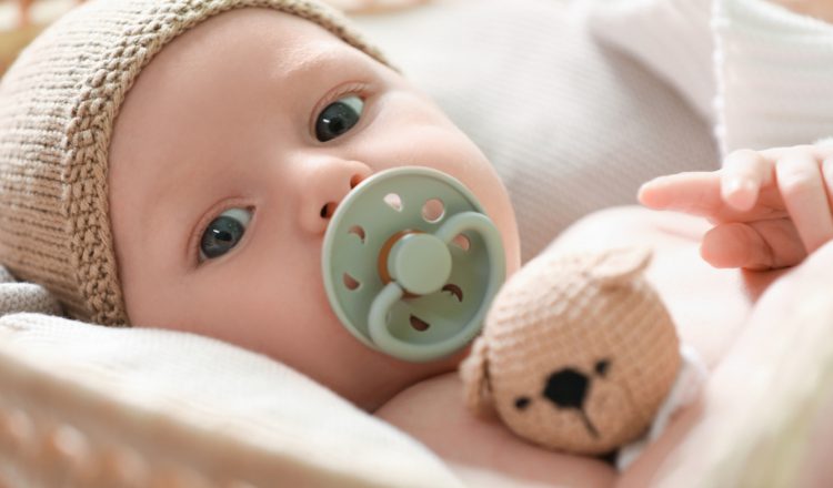 Imagem do rostinho de um bebê. Ele está deitado, tem a pele clara e os olhos castanhos. Está usando uma chupeta verde oliva e uma touca tricô cru. Ele está segurando um ursinho também feito de tricô.