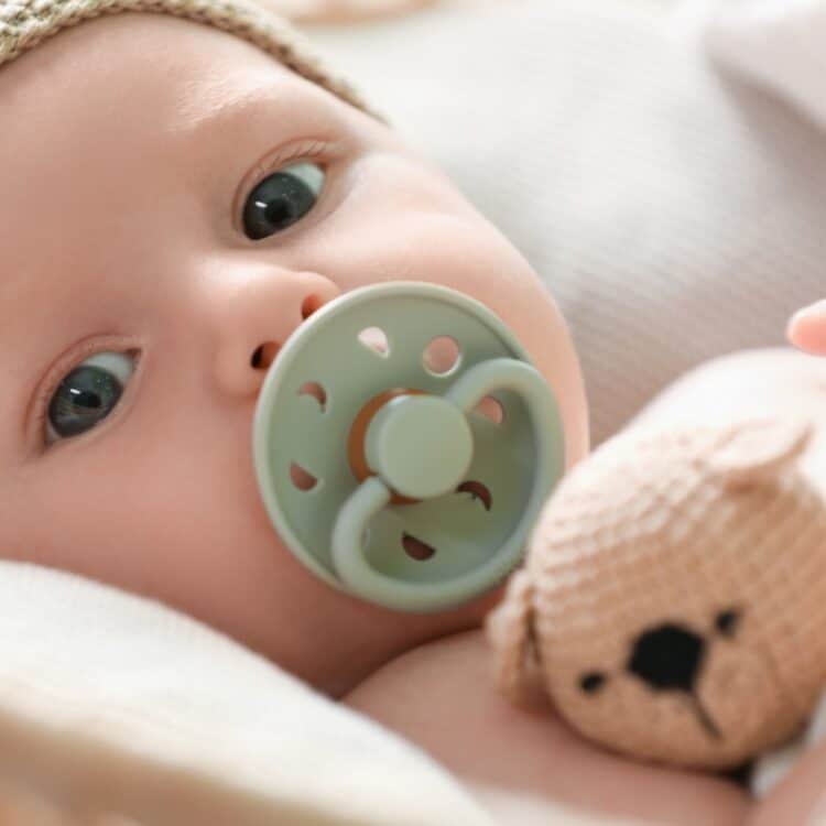 Imagem do rostinho de um bebê. Ele está deitado, tem a pele clara e os olhos castanhos. Está usando uma chupeta verde oliva e uma touca tricô cru. Ele está segurando um ursinho também feito de tricô.
