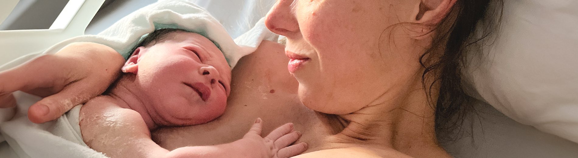 Imagem de mãe e bebê logo após o parto. A mãe está deitada em contato pele a pele com o bebê, que está de bruços sobre o seu peito. Eles estão olhando um para o outro.