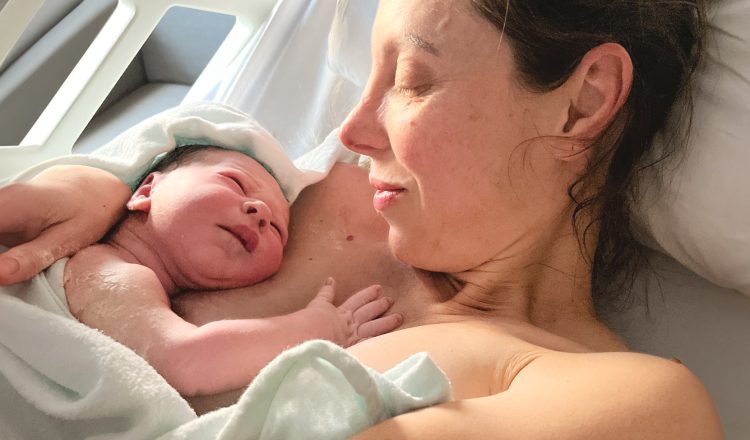 Imagem de mãe e bebê logo após o parto. A mãe está deitada em contato pele a pele com o bebê, que está de bruços sobre o seu peito. Eles estão olhando um para o outro.