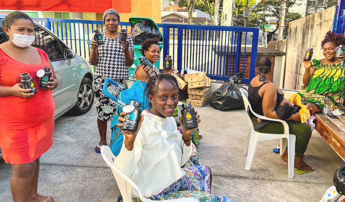 Na imagem estão seis mulheres, gestantes atendidas pelo projeto Be Generous em 2023. Elas estão mostrando os suplementos recebidos por meio do projeto. Quatro estão sentadas e duas estão em pé. Todas estão felizes com os suplementos recebidos.