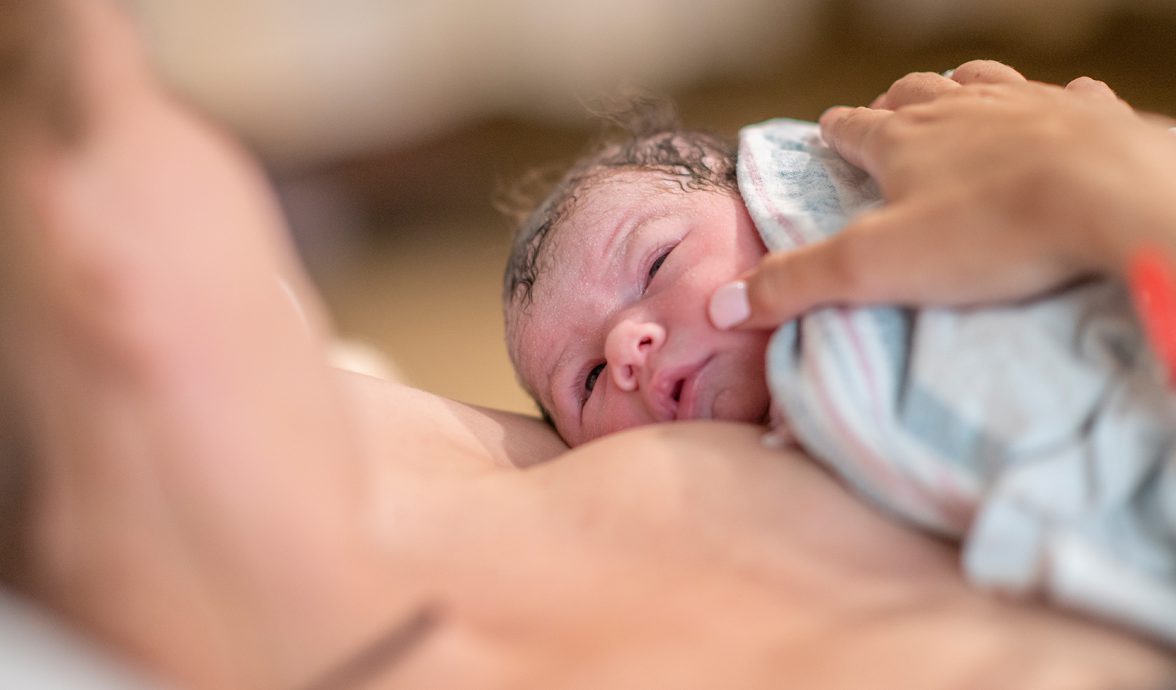 Imagem de um bebê recém-nascido na golden hour. Ele está sobre o peito da mãe, em contato pele a pele. Ambos estão se olhando. O bebê tem a pele clara e o cabelo escuro.