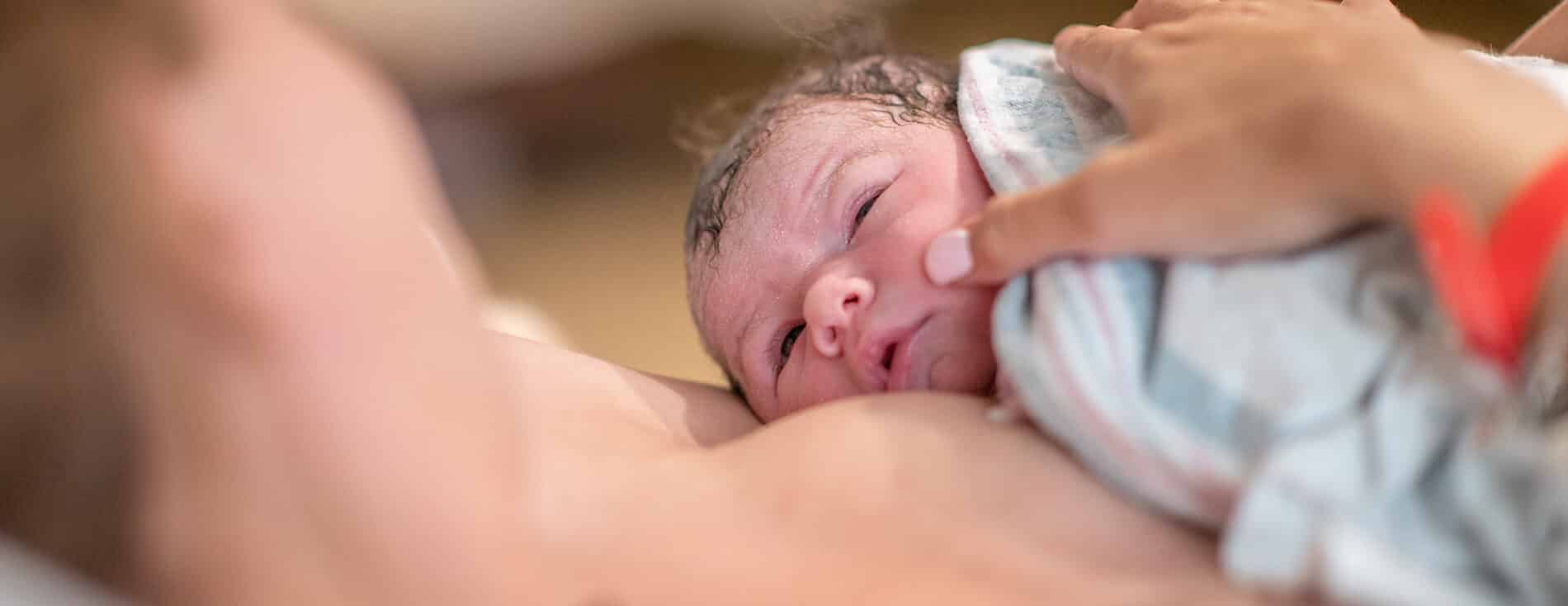 Imagem de um bebê recém-nascido na golden hour. Ele está sobre o peito da mãe, em contato pele a pele. Ambos estão se olhando. O bebê tem a pele clara e o cabelo escuro.
