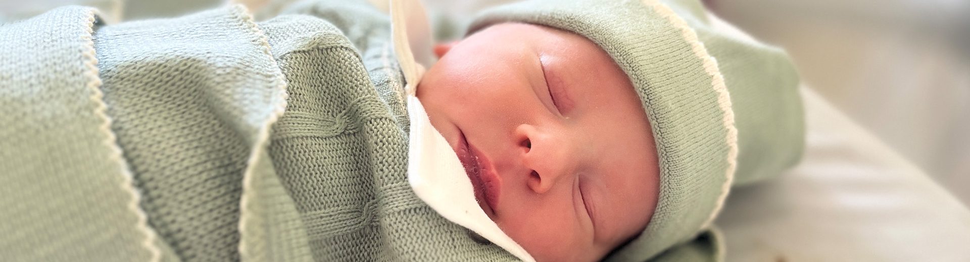 Imagem de um bebê recém-nascido. Ele tem a pele branca e os olhinhos puxados. Está dormindo em um bercinho e veste uma roupa de linho verde oliva, com detalhes em bege. Veste touca e está coberto com uma matinha do mesmo tecido e cor.