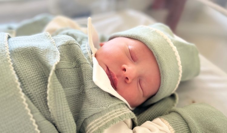 Imagem de um bebê recém-nascido. Ele tem a pele branca e os olhinhos puxados. Está dormindo em um bercinho e veste uma roupa de linho verde oliva, com detalhes em bege. Veste touca e está coberto com uma matinha do mesmo tecido e cor.