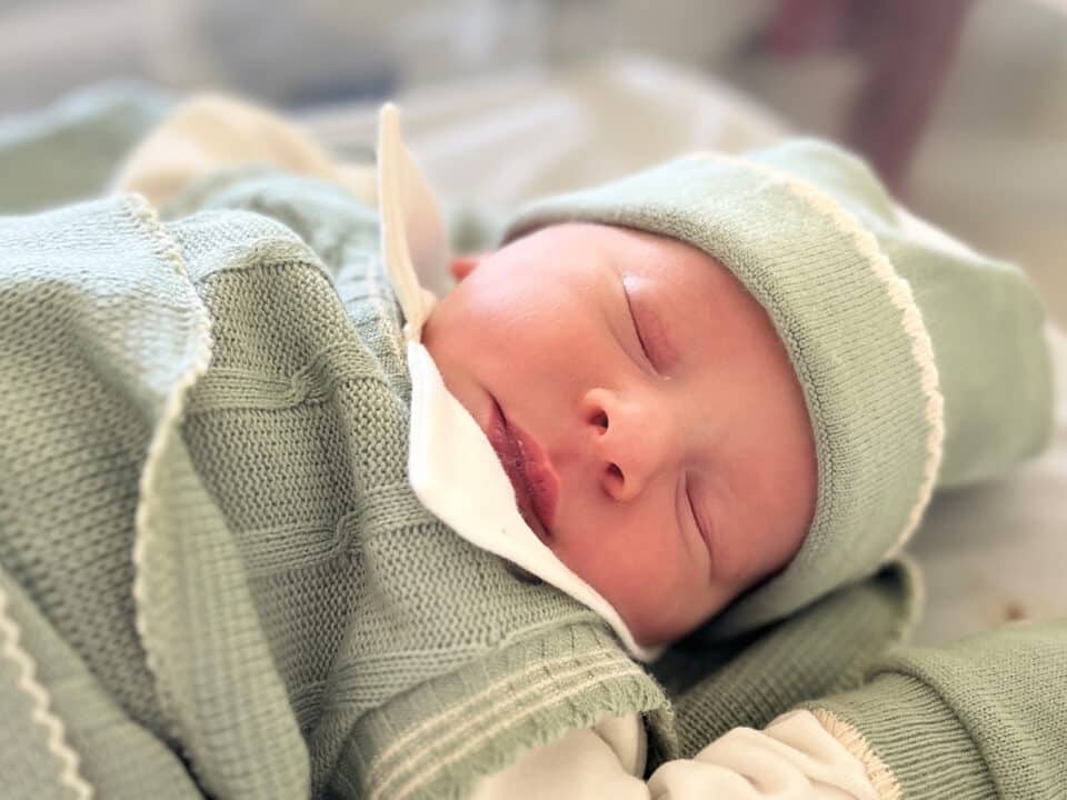 Imagem de um bebê recém-nascido. Ele tem a pele branca e os olhinhos puxados. Está dormindo em um bercinho e veste uma roupa de linho verde oliva, com detalhes em bege. Veste touca e está coberto com uma matinha do mesmo tecido e cor.