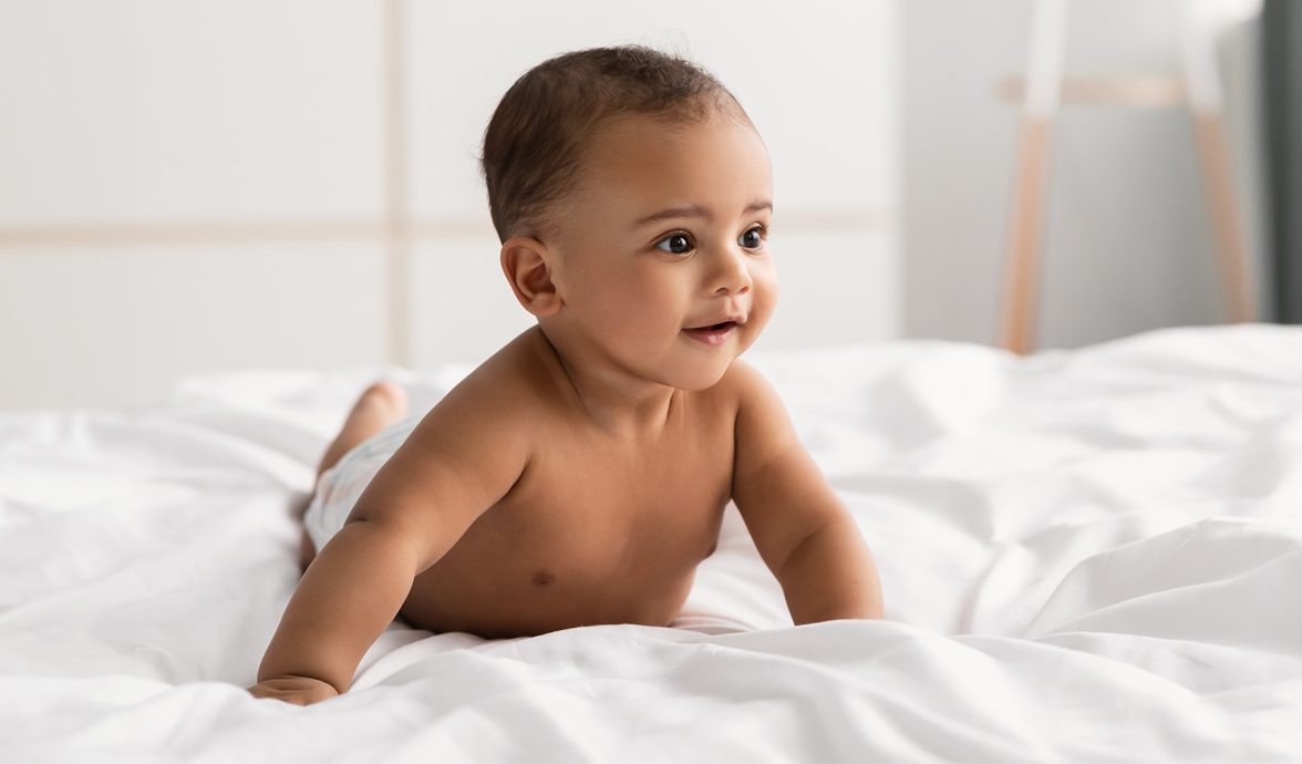 Imagem de um bebê de bruços em cima de um lençol branco, em cima da cama. Ele está apenas de fralda, tem a pele morena, olhos e cabelos escuros e está sorrindo.