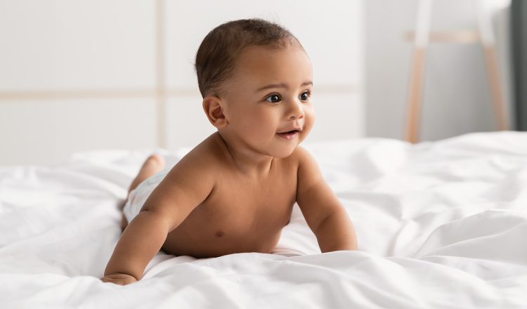 Imagem de um bebê de bruços em cima de um lençol branco, em cima da cama. Ele está apenas de fralda, tem a pele morena, olhos e cabelos escuros e está sorrindo.